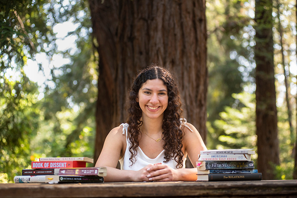 Photo of young woman with redwood trees behind her and stacks of books in front of her.
