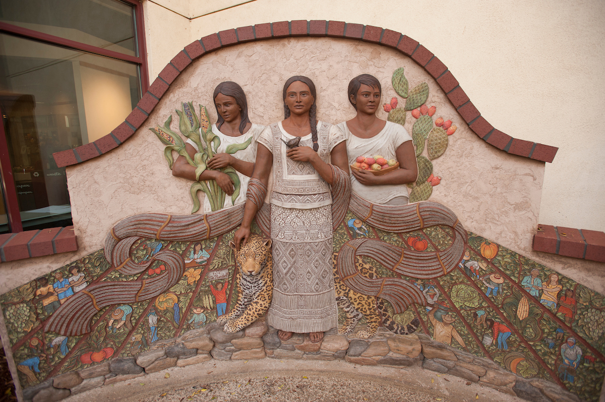 A bas relief sculpture with three Latina women: one on left holding corn, one on holding basket of cactus fruit, one in center with a bird on one hand and petting a leopard with other hand