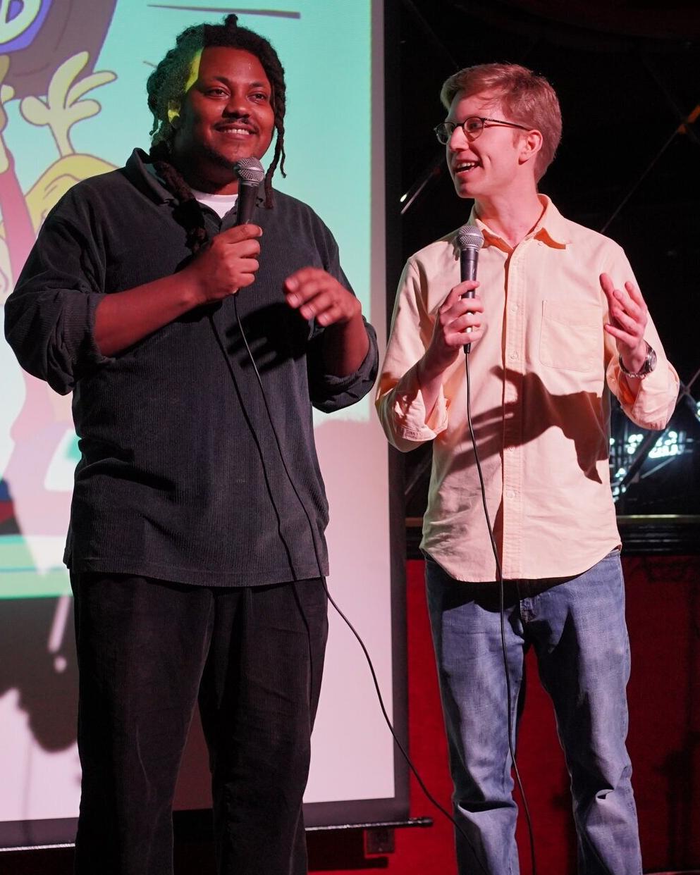 Two comedians, one black and one white, holding microphones, smiling