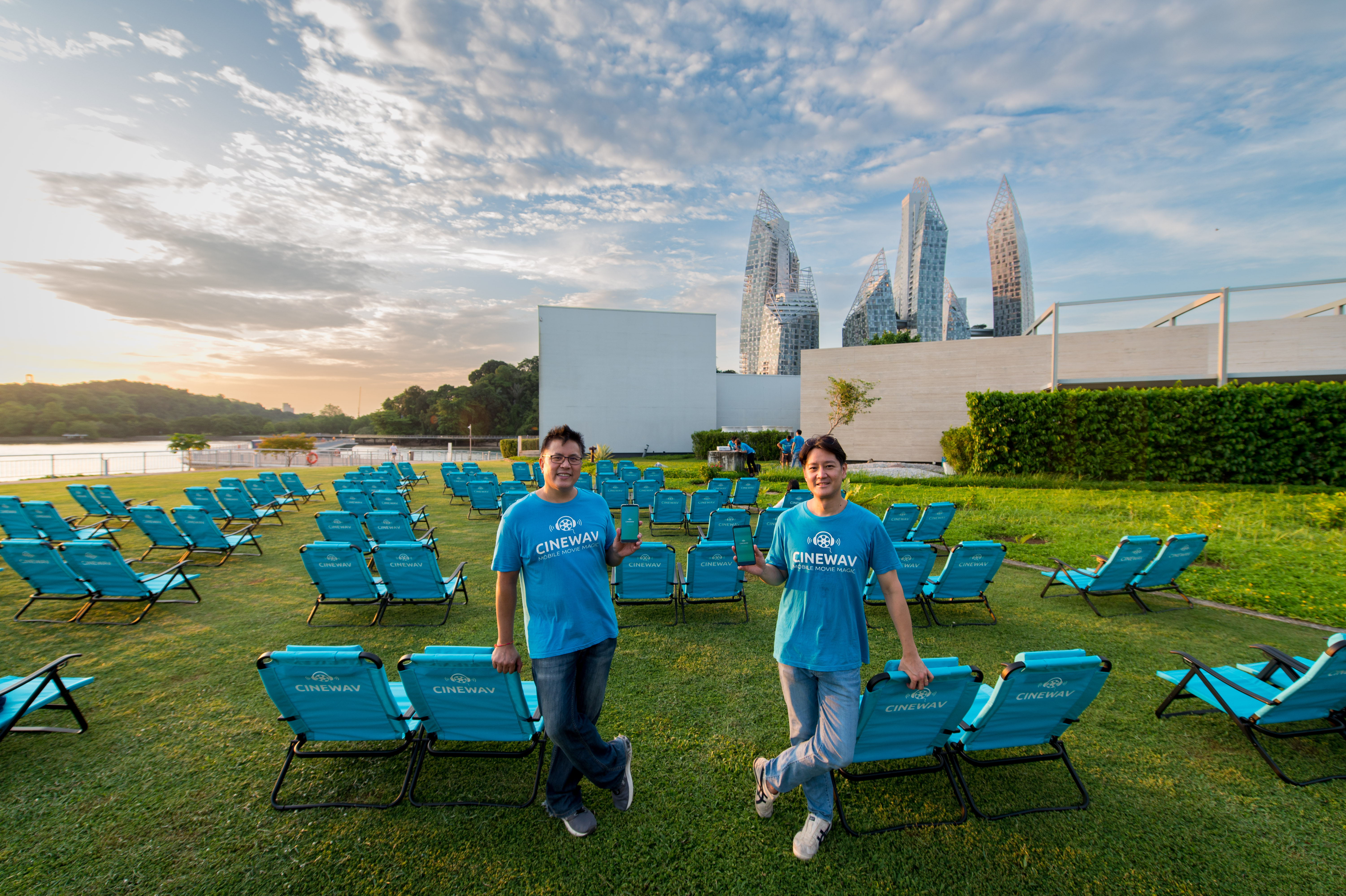 Two smiling men in blue Cinewav t-shirts hold up phones with the app at an outdoor pop-up cinema on a green lawn