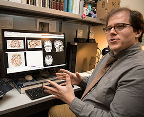 UC Davis neuroscience researcher turning in his chair to talk with someone off camera, his computer screen showing images of brain scans.