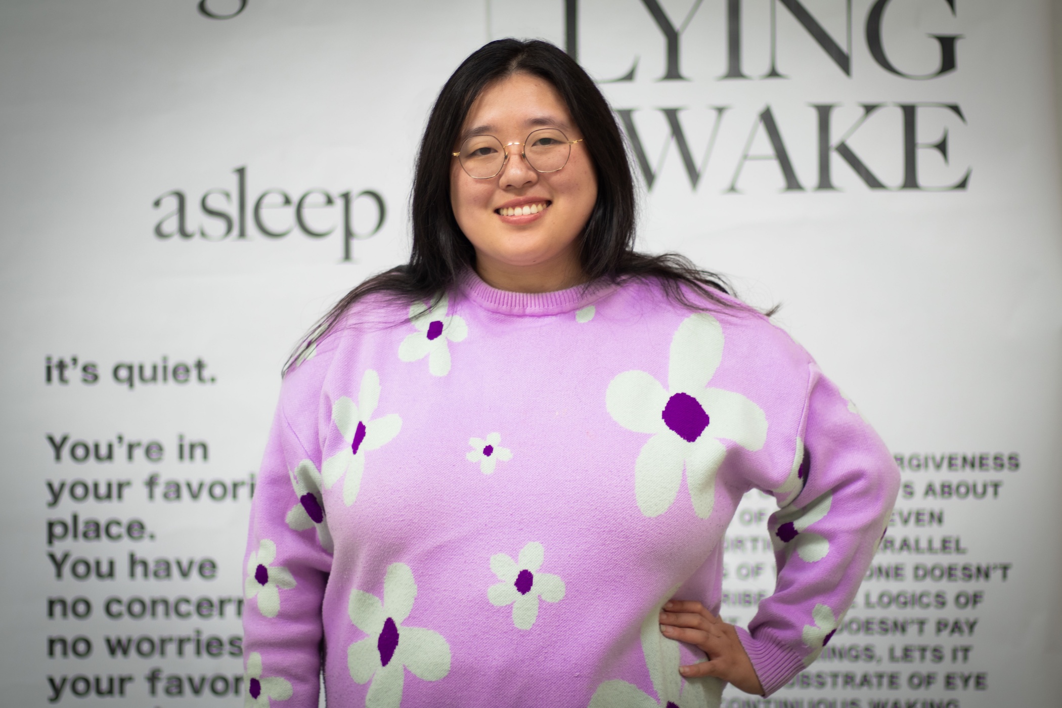 woman with glasses and long dark hair in lilac sweater with abstract flower designs standing in front of large text on wall