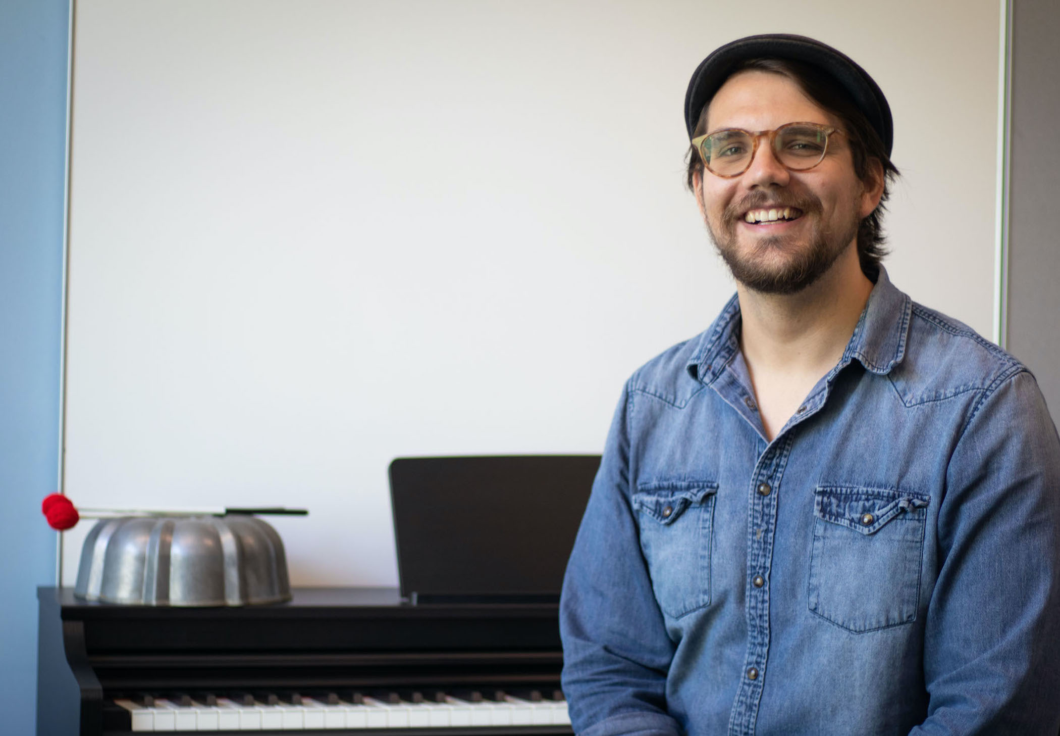 Man sitting to the right side of photo, wearing bark cap, blue denim shirt, glasses, smiling, in front of a keyboard