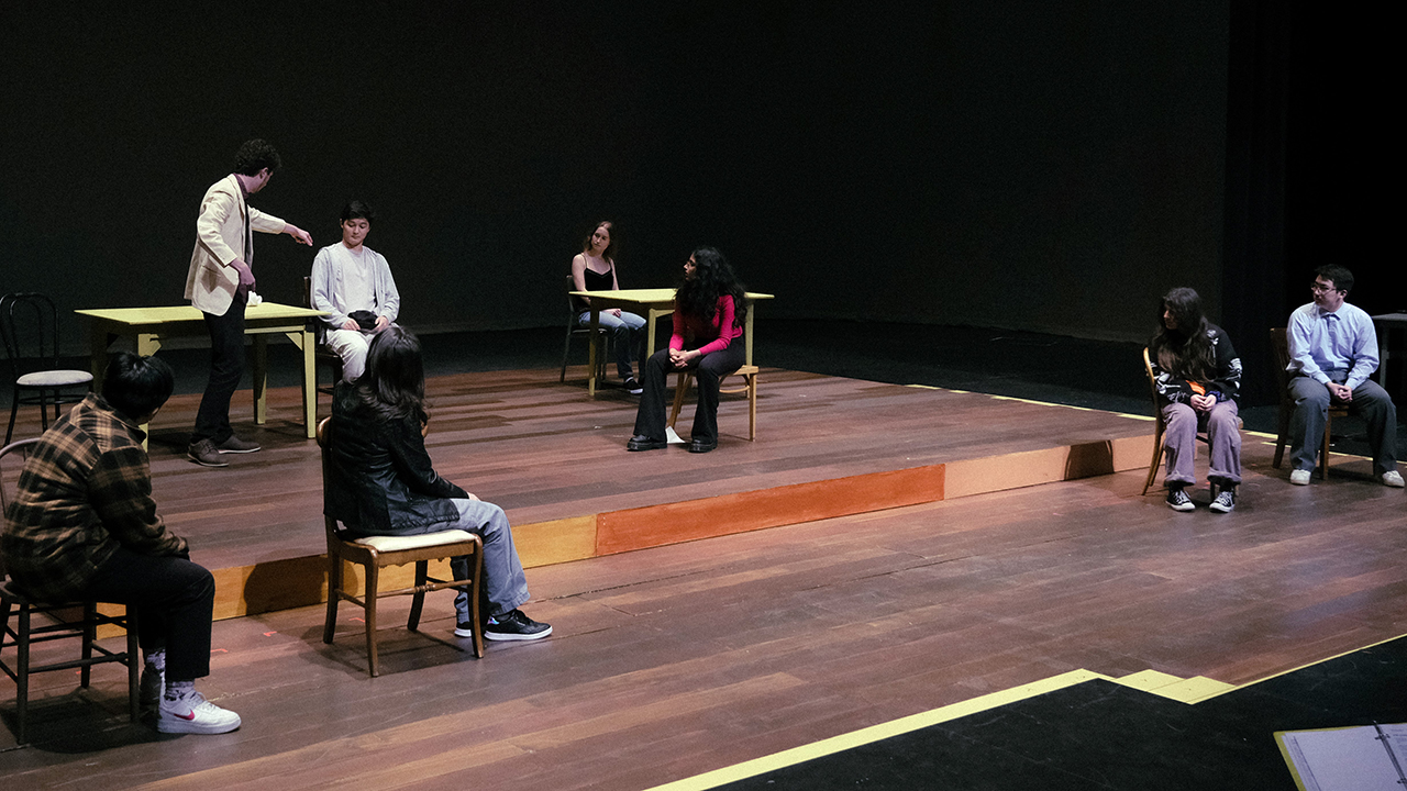 Eight undergraduate students rehearse The Laramie Project on a 2-level stage, scattered around small tables or seated on chairs
