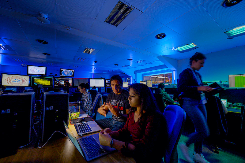 UC Davis physics researchers in a control room at CERN filled with computers and illuminated with blue lighting.