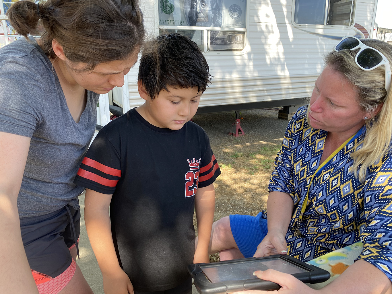 A mother and her third-grader son standing and looking at a Chromebook held by seated teacher, outside their mobile home.  