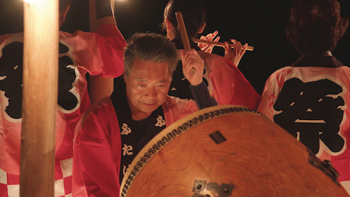 photo of a man playing large drum with other musicians in the background. Overall color is dark orange and brown. 