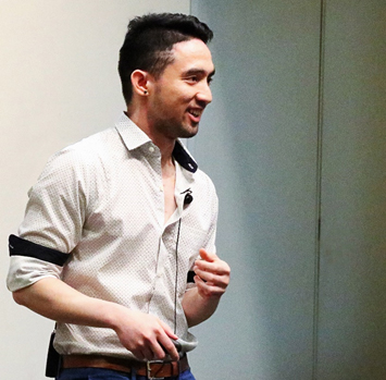 Young man with dark heard, standing with microphone clipped to his dress shirt, sleeves rolled up