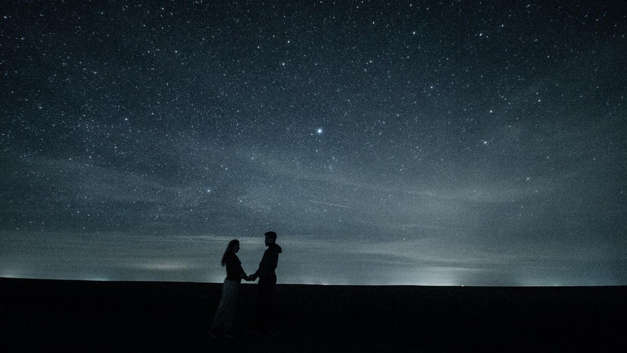 Photo: Couple facing each other, holding hands, with starry night sky in background. 