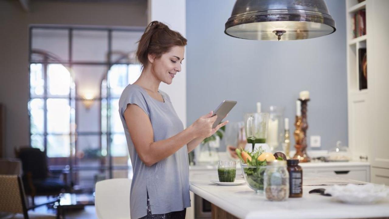 Woman standing in kitchen and looking at a tablet. 