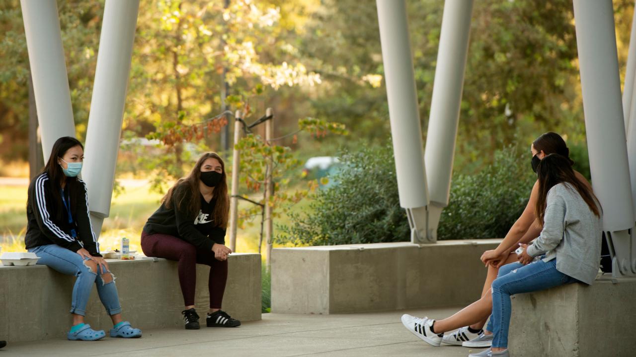 Four female students in masks, sitting on concrete benches outside.