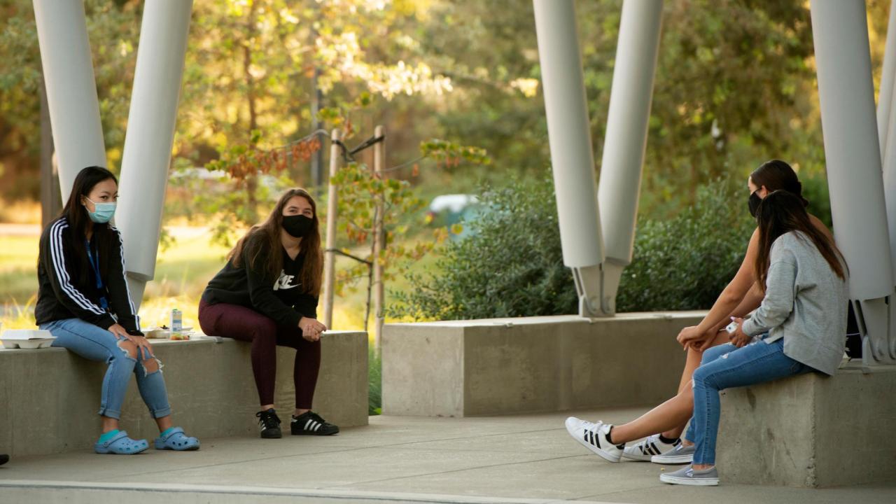 Four female students in masks, sitting on concrete benches outside.