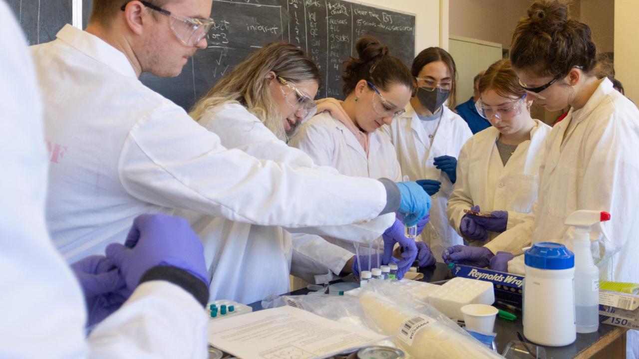 A group of students in lab attire gather around a table to investigate aromas associated with whiskey.