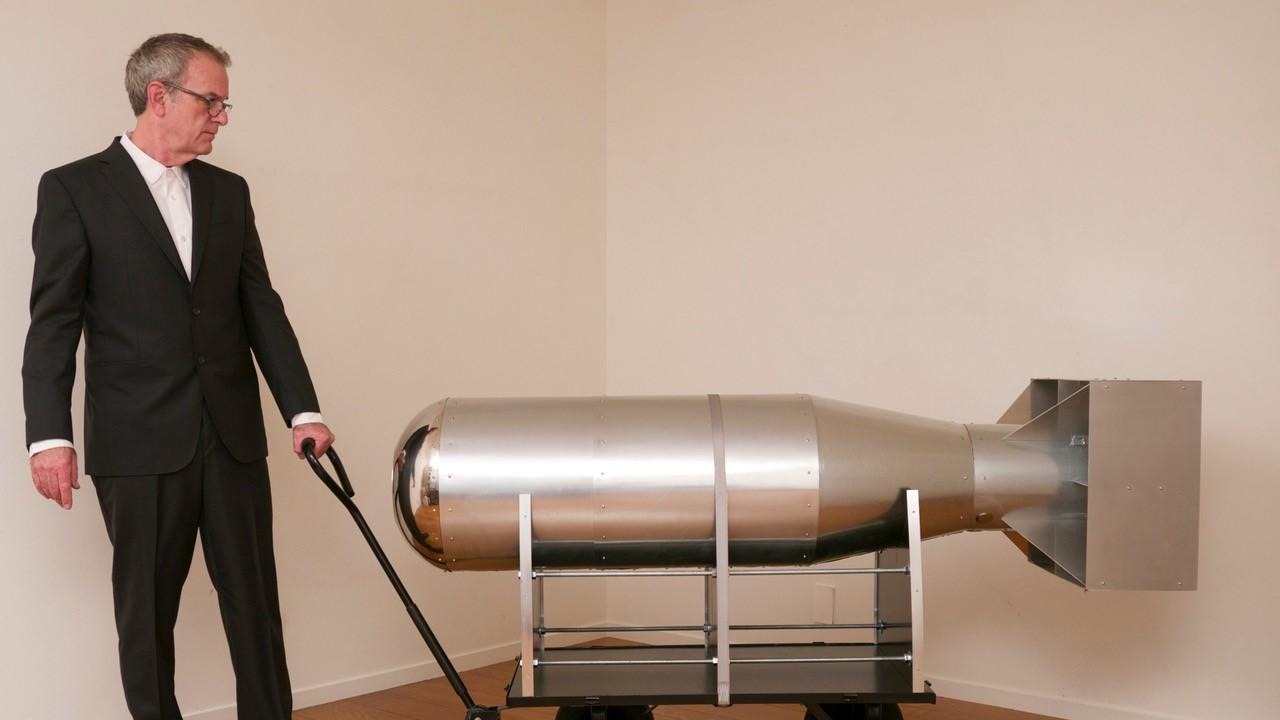 Stephen Whisler, dressed in a suit, pulls a replica of a bomb on a cart 