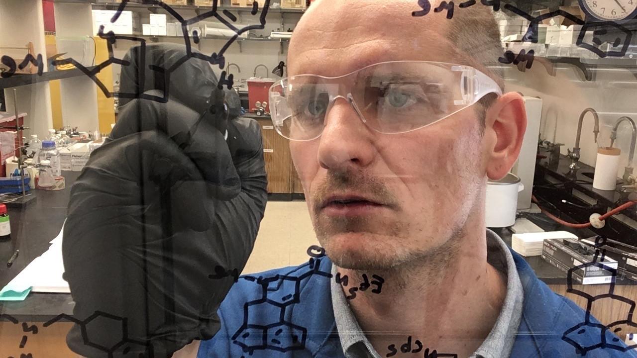 Close-up of David Olson in his lab wearing safety glasses and gloves as he draws on a clear plexiglass board.