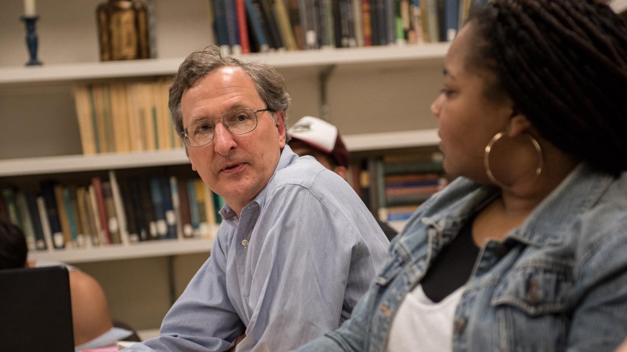 man with glasses turning toward a woman with long dark hair bookshelves in the backgroun