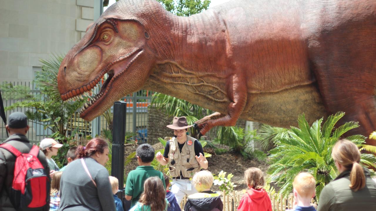 Benjamin Faulkner is speaking to a large crowd of people as he stands in front of a large dinosaur replica