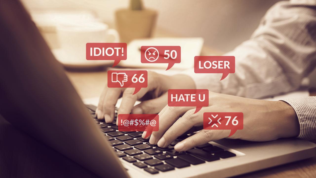 Hands on a laptop keyboard with red thought bubbles with words "Idiot!" "Hate U" and "Loser," icons for "Don't Like" and symbols representing expletives.