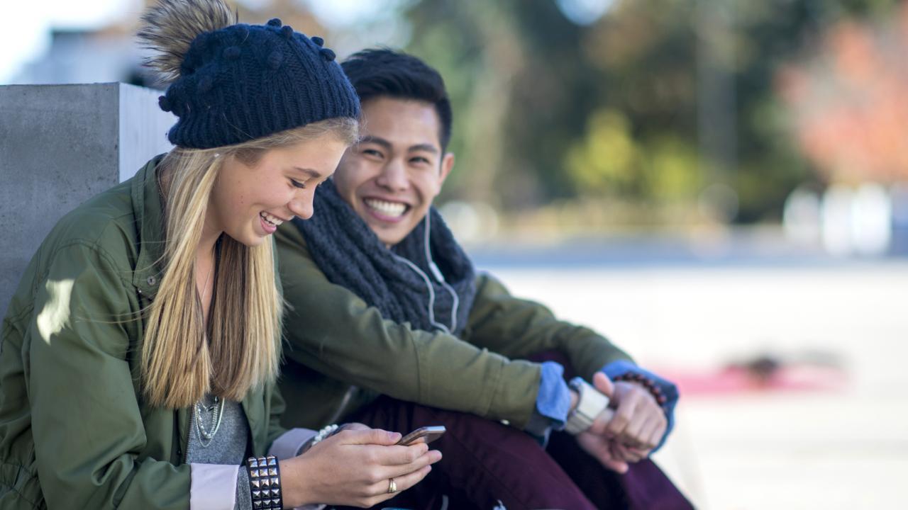 Two students are on their phone, chatting and smiling. 