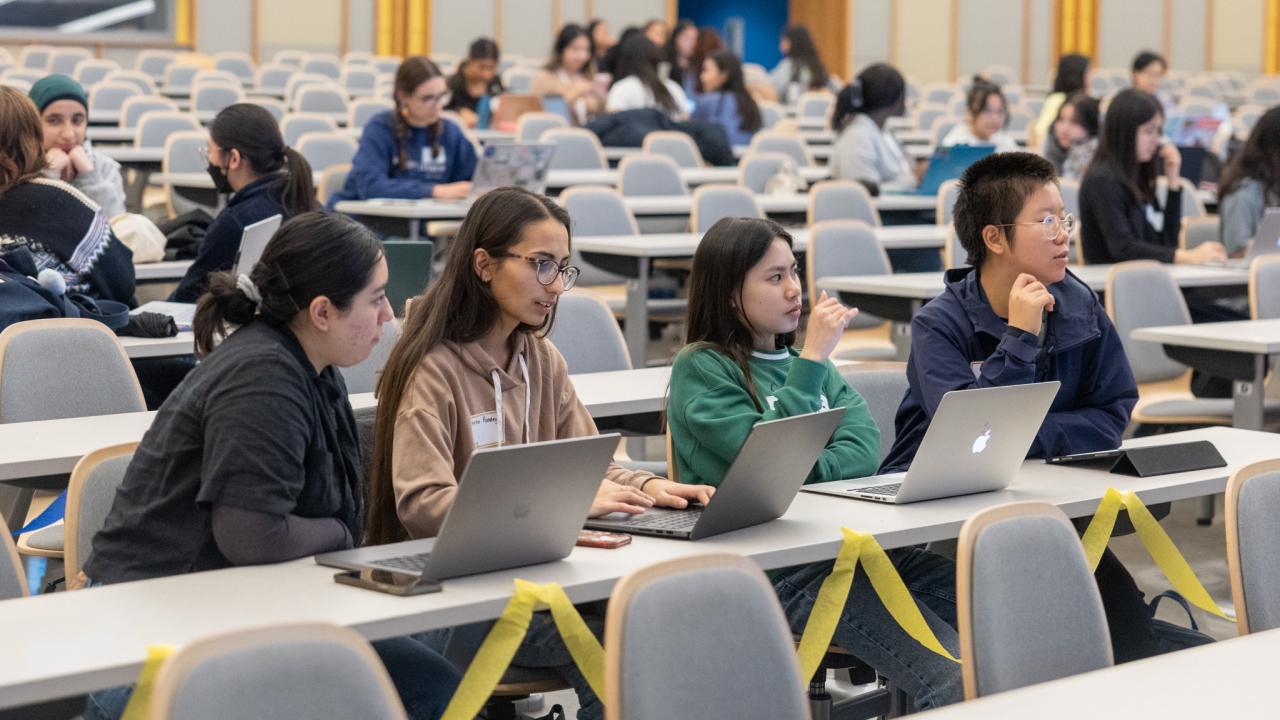 Four girls are sitting behind their laptop computers in a large lecture hall