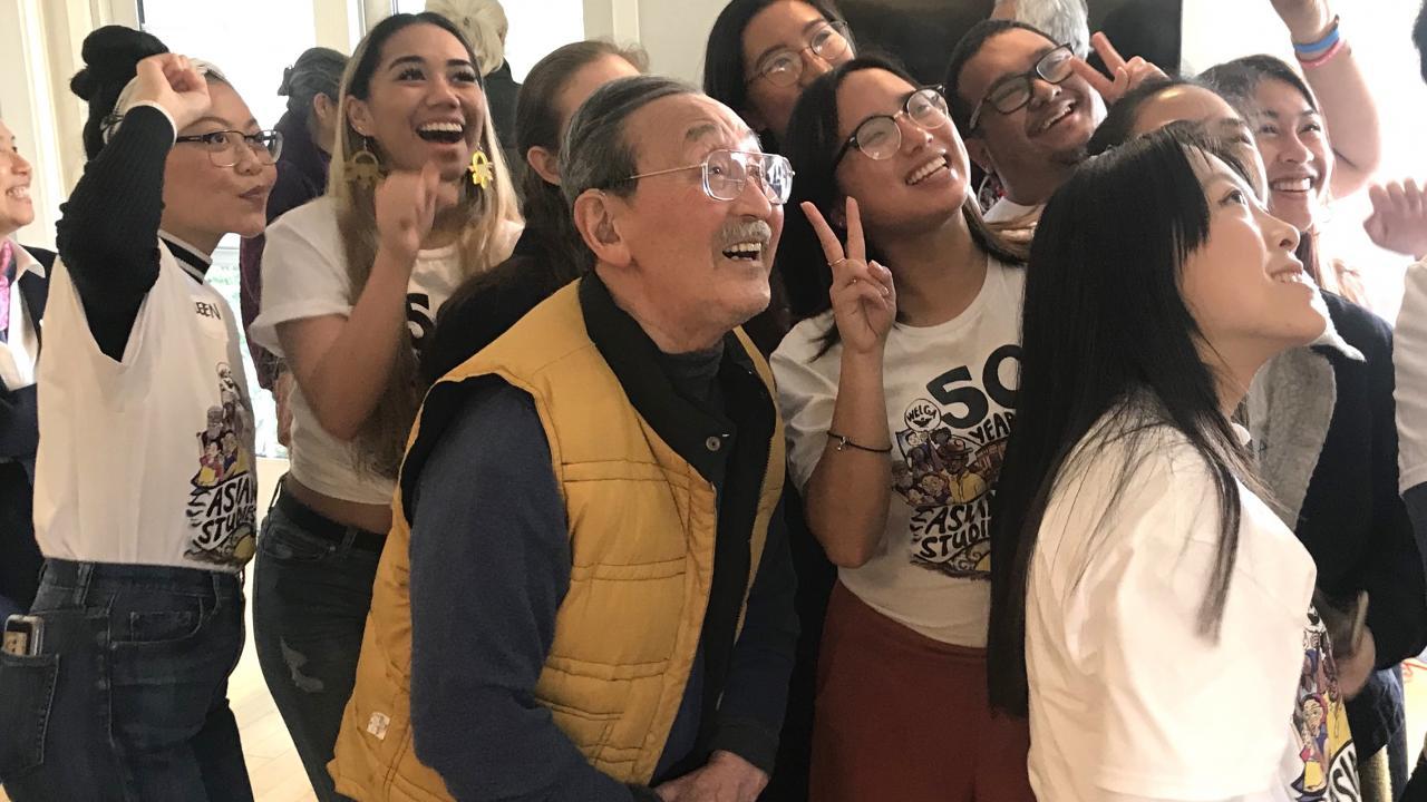 Older man in gold colored vest and glasses posing with group of young people all facing right 