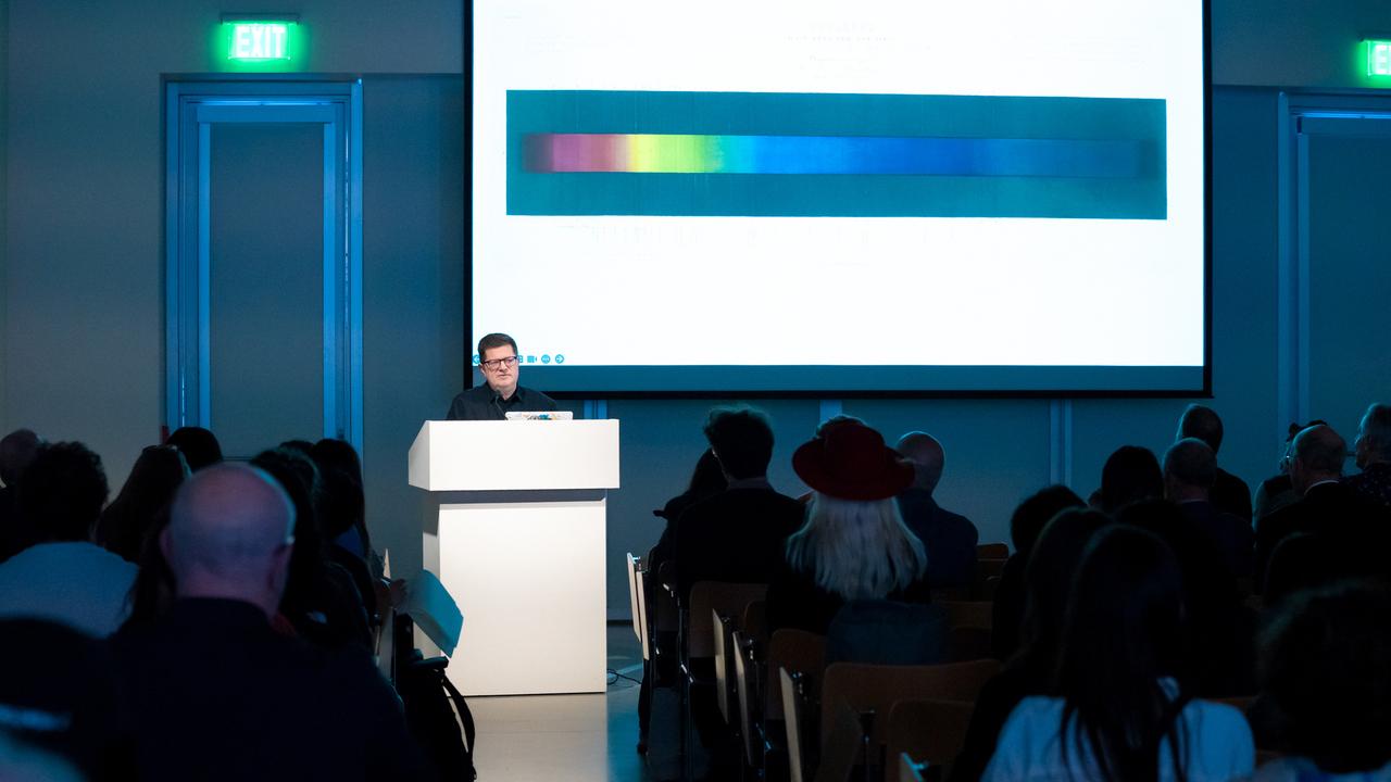 Josiah McElheny at podium with presentation slide featuring a horizontal multi-colored strip