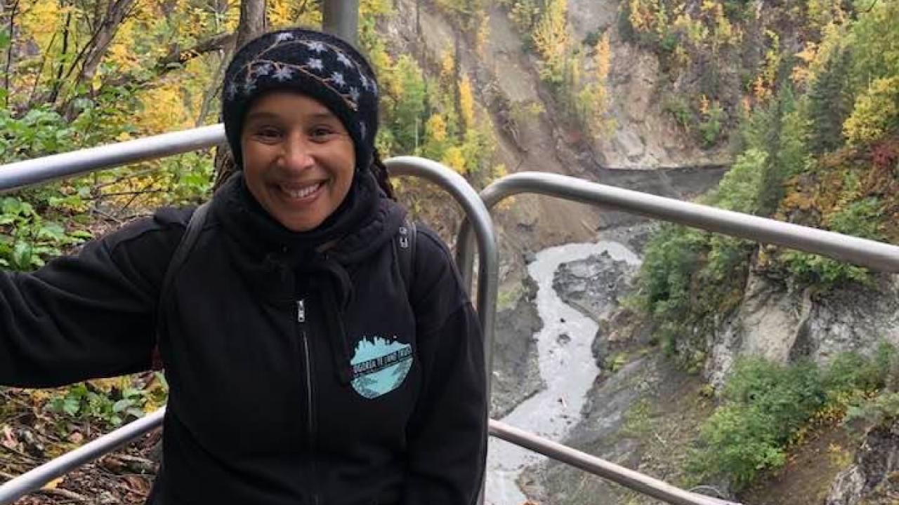 Woman dressed in heavy clothing, crouching down and smiling, in background and below river and forest 