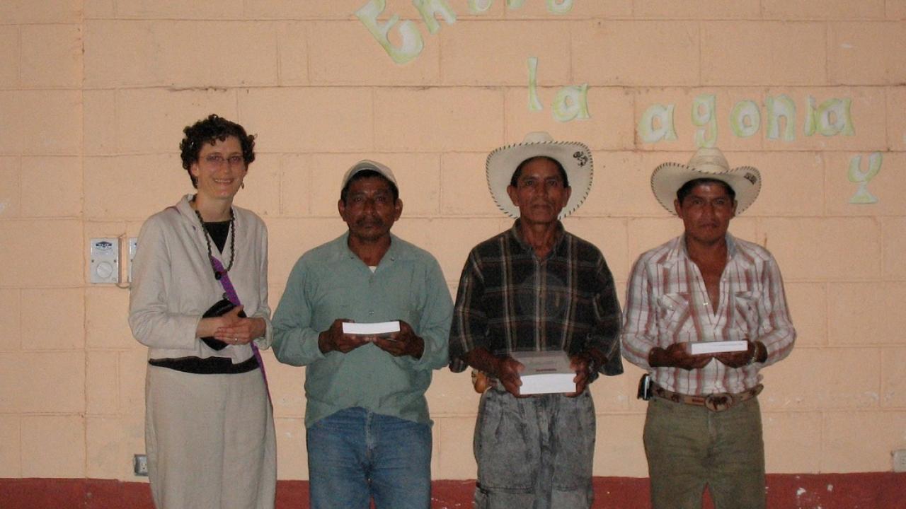 group of four people standing in a line, one woman on the left wearing loose gray jack and pants and three men, two in cowboy hats, one in cap in work clothing each holding a book  