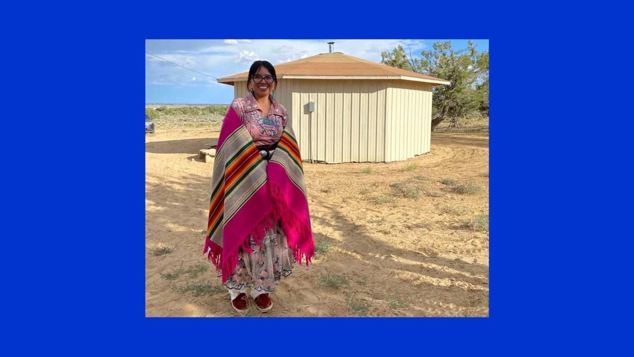 woman wearing brightly colored wrap over rose colored floral dress. Wearing thick framed black glasses, black hair tied back. Standing in from of a small off white wooden buuilding