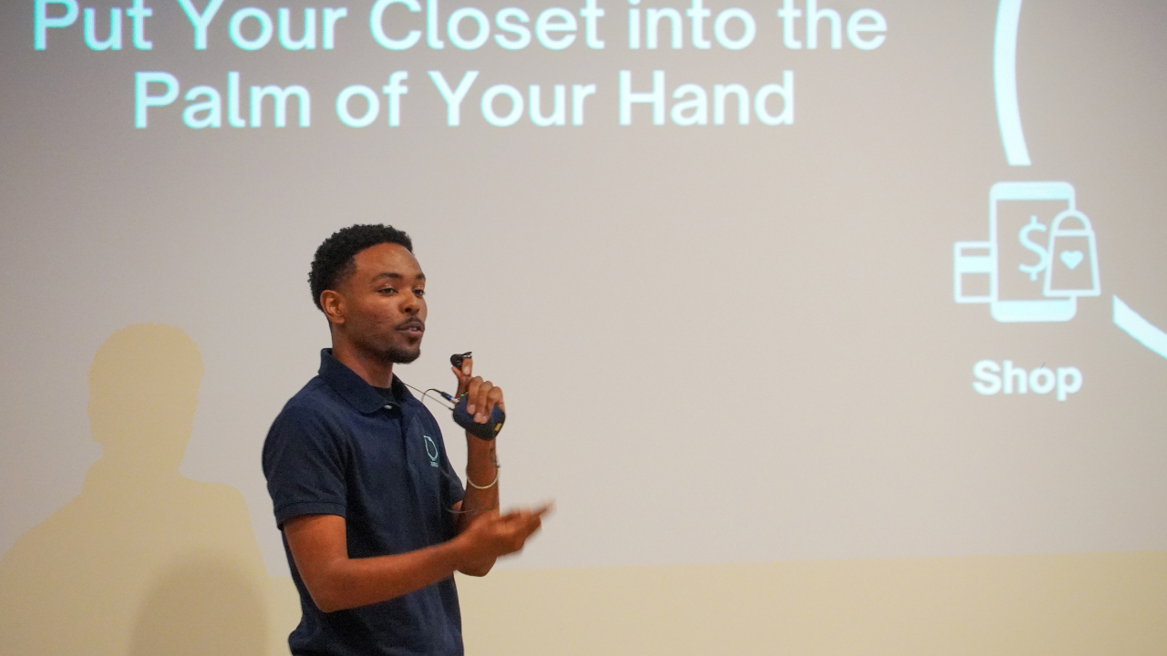 Young Black man in navy blue polo shirt, speaks into small handheld microphone. Slide presentation behind him reads, "Put Your Closet Into the Palm of Your Hand." To right, part of a circle shows with icons for money and a shopping bag and the word "Shop.”