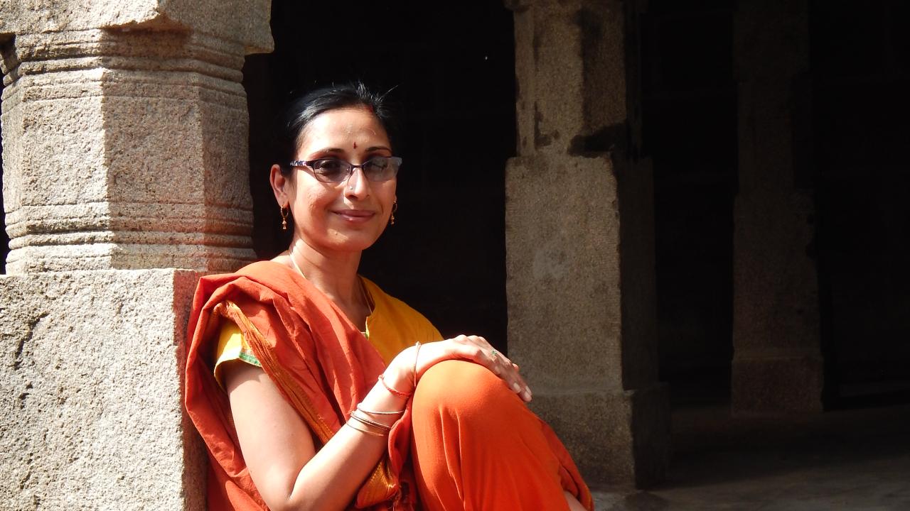 Woman with dark hair and glasses, dressed in saffron colored sari leaning against a stone pillar.