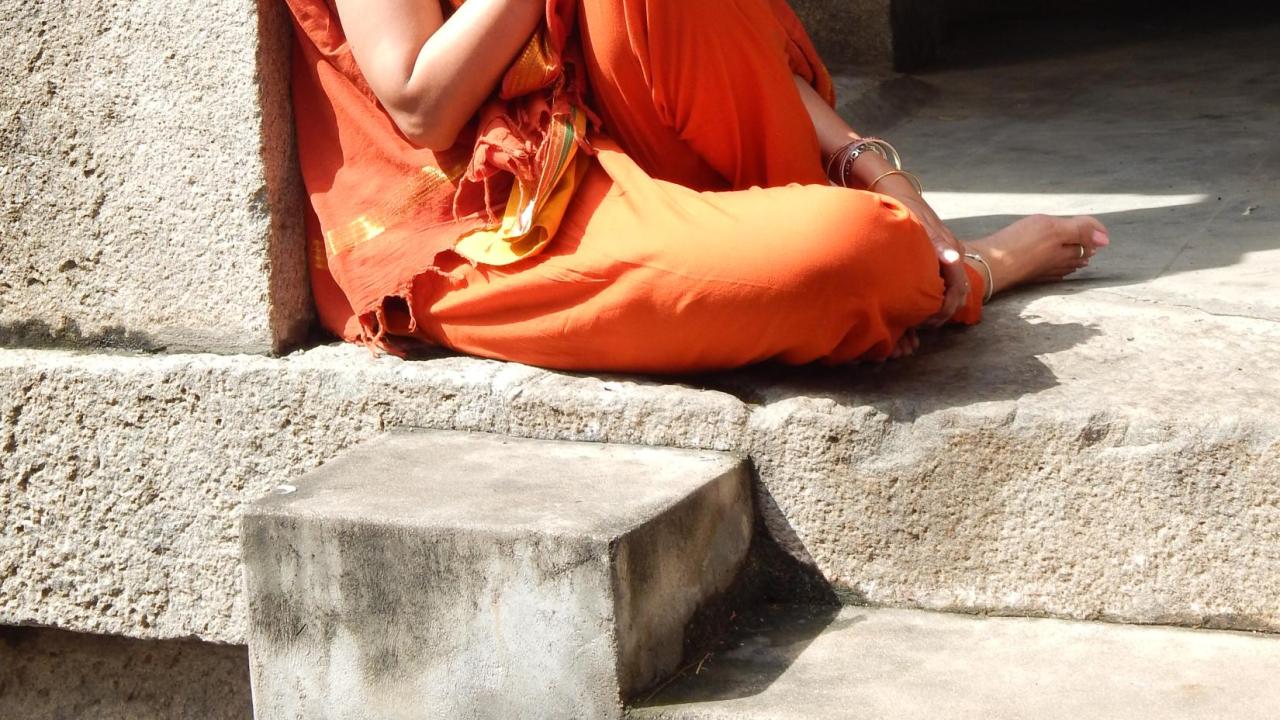 Woman with dark hair and glasses, dressed in saffron colored sari leaning against a stone pillar.