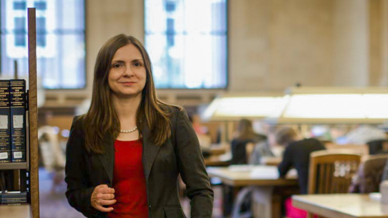 UC Davis psychologist standing in the spacious library reading room. Behind her are large windows and students studying at desks with brass and glass reading lamps.