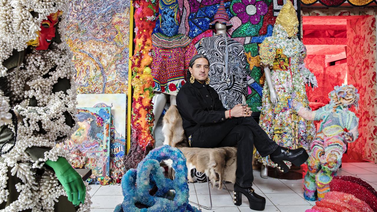 Photo of man with dark hair and black clothing sitting in a chair surrounded by art that is brightly colored 