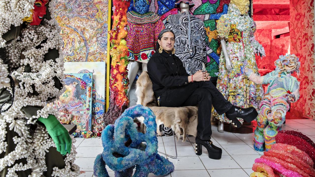 Photo of man with dark hair and black clothing sitting in a chair surrounded by art that is brightly colored 