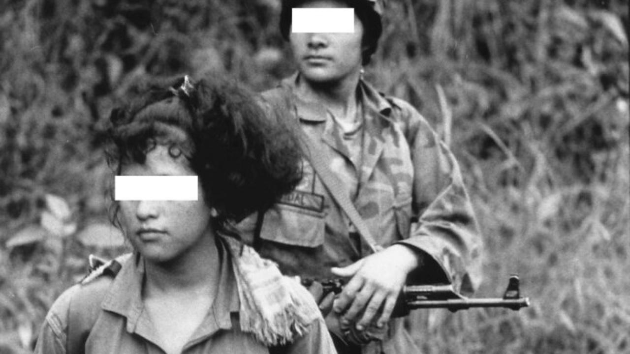 black and white photo of two women their eyes blanked out by white bars in military fatigues holding guns.