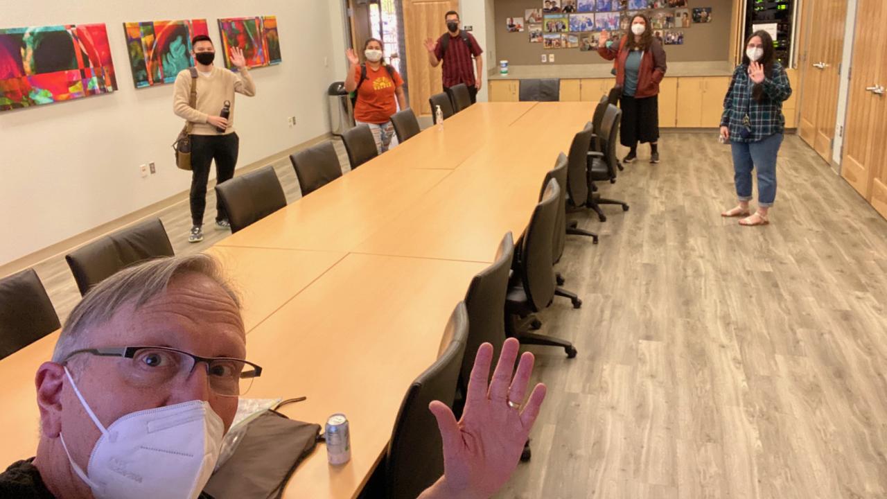 A professor up close and five students standing at other end of a conference table wave at the camer