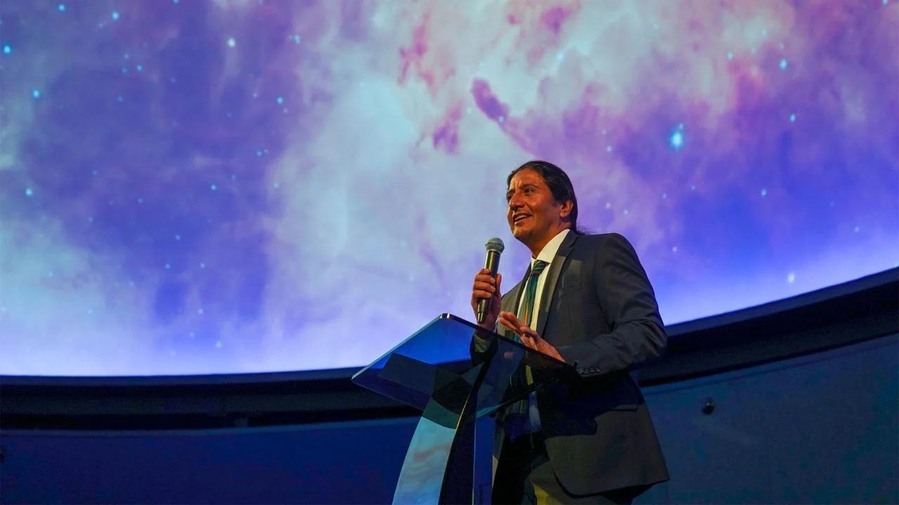 Manuel Calderón de la Barca Sánchez is holding a microphone and speaking in front of a large display of the universe behind him