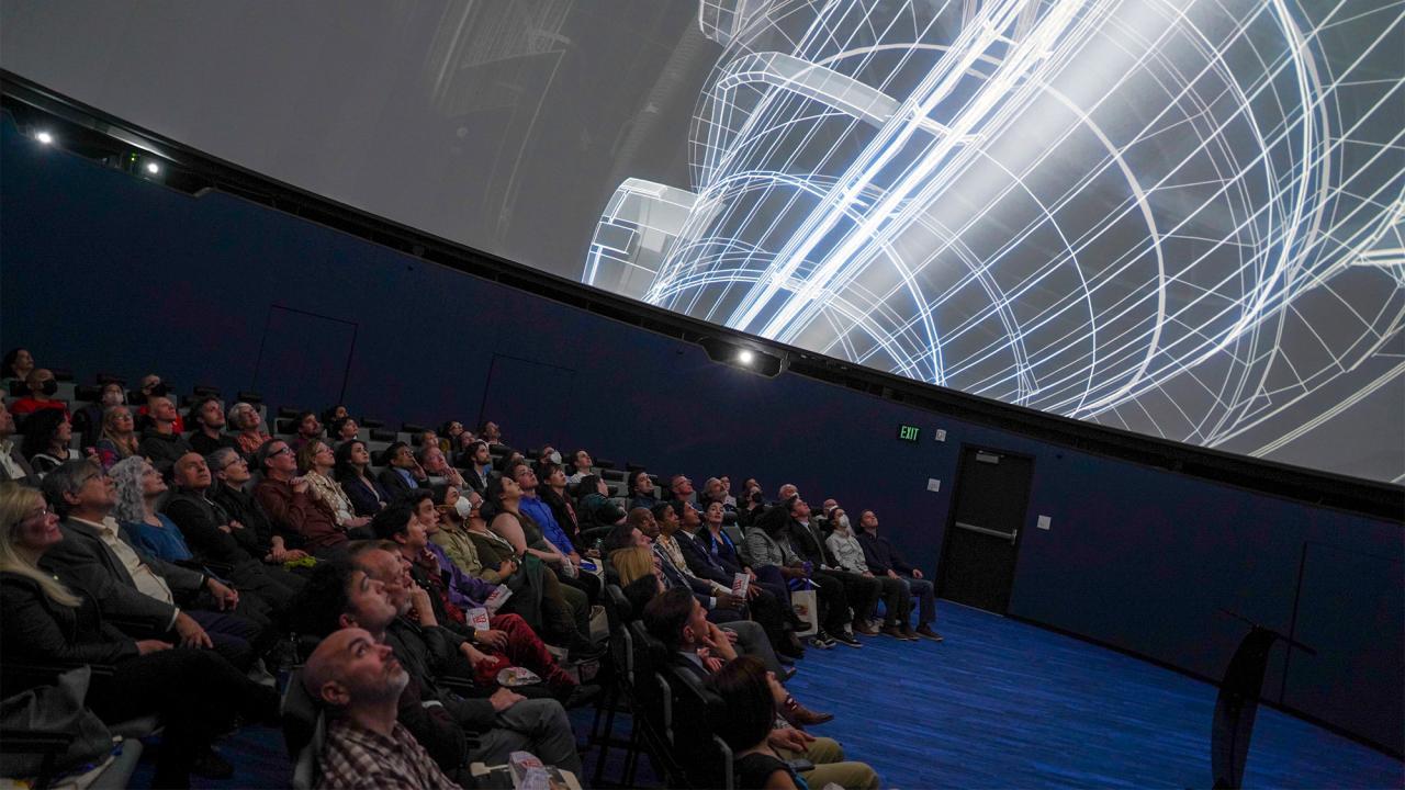 About 100 people sitting in a theater looking upwards at a planeterium screen.