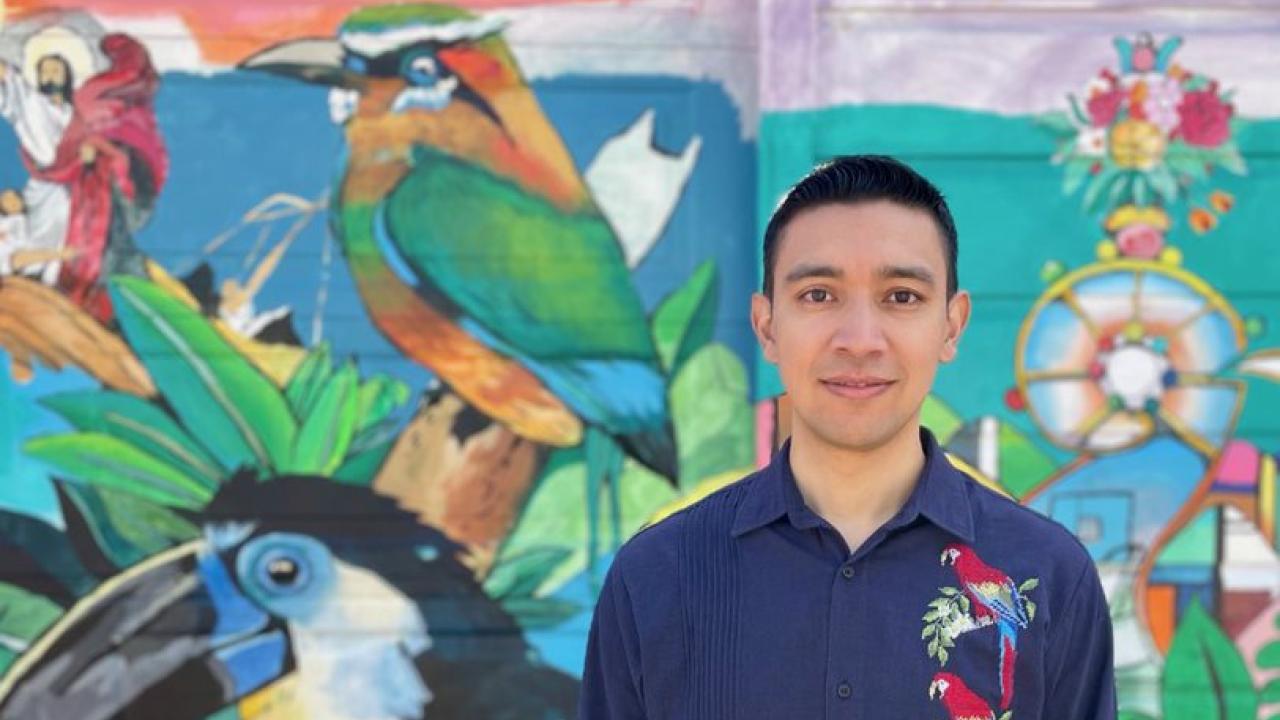 man in dark blue shirt with colorful embroidery on the left side, short dark hair looking directly at the camera in fronnt of a colorful mural with people, green space and mountain. 