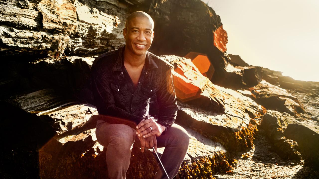 Man sitting on rocky beach, wearing dark shirt and brown pants, smiling at the camera and holding a conductor's baton 