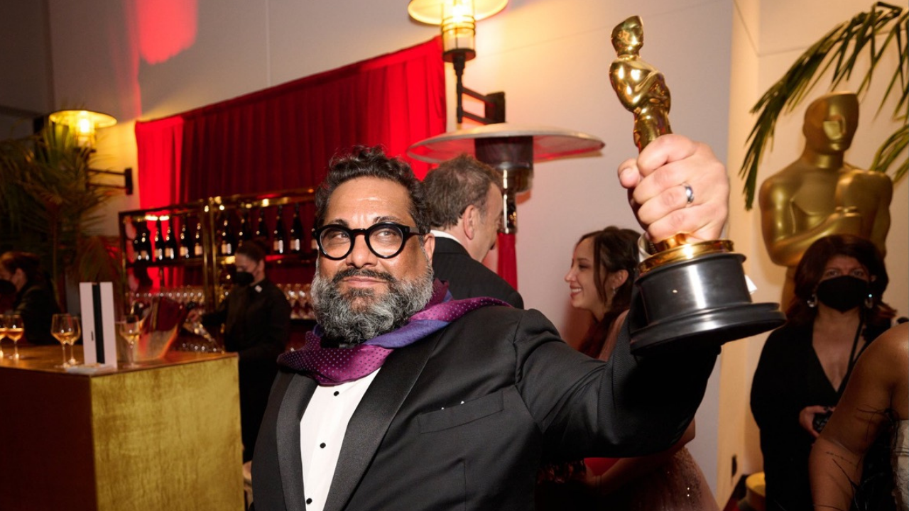 Bearded UC Davis alumnus dressed in tuxedo and a colorful scarf around his neck, holds up his Oscar backstage at the Academy Awards