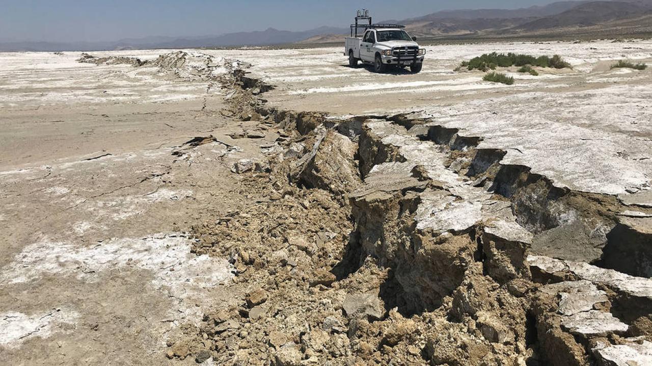 A fault scarp from the Ridgecrest earthquake cuts across a dry lake bed with a white truck in the background.