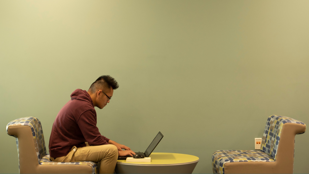 A male Asian student sitting in student lounge, typing on laptop