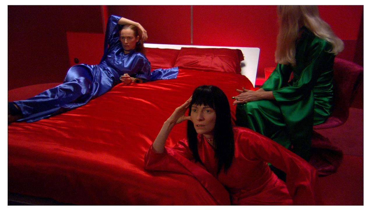 Image of three women, all the actor Tilda Swinton, on a red bed. ONe on left wearing blue gown, center green, right  red