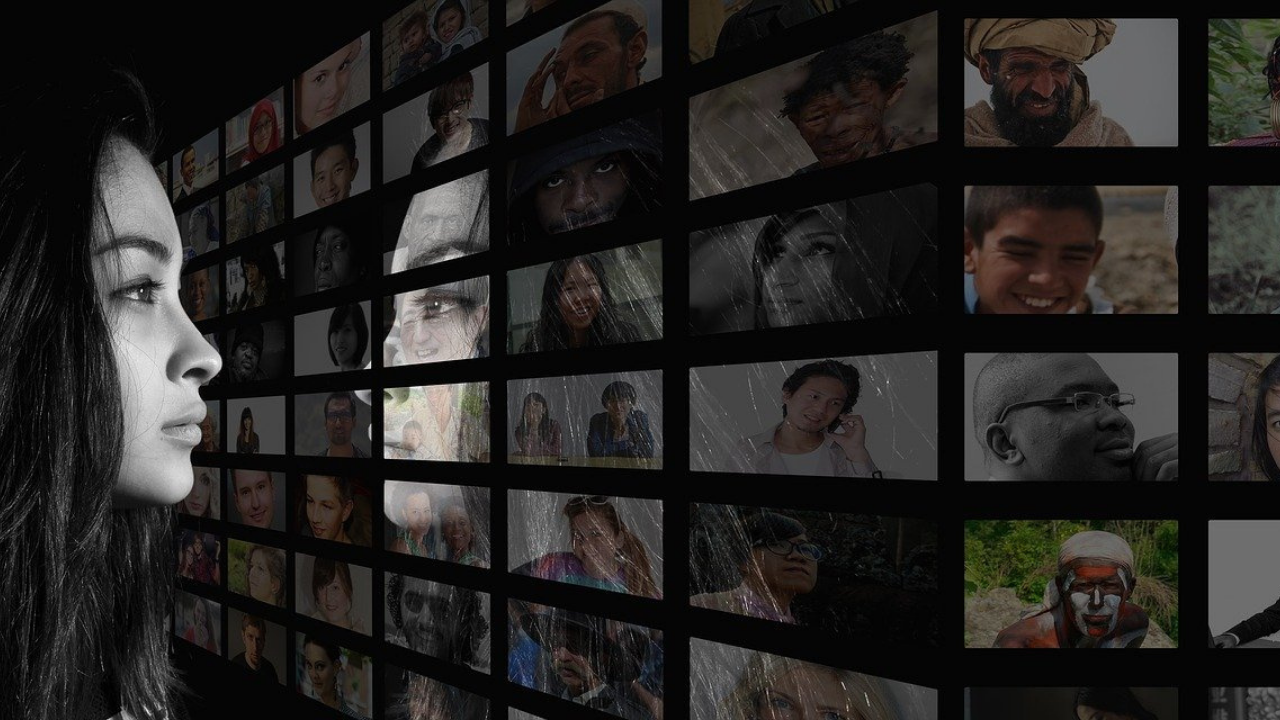 Woman looking into, and reflected in, a large screen of a large screen with a grid of multiple photos of globally diverse faces.