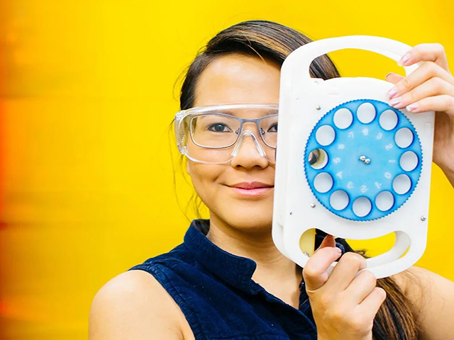 Asian female student wearing clear safety glasses over prescription glasses, holding a white & blue eye prescription lens checker tool next to her face