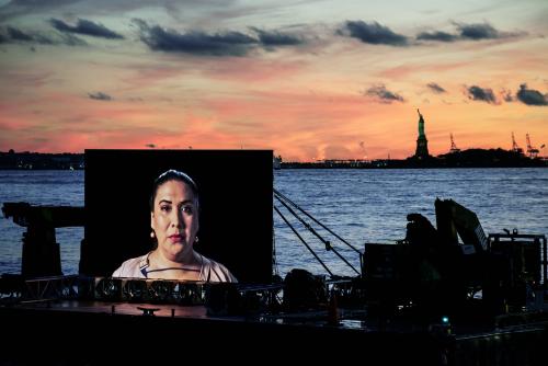 image of a barge on the water close to shore. On a screen on the barge is a close up photo of a woman with dark hair. in the background is a colorful sunset and the south end of New York and the Statue of Liberty
