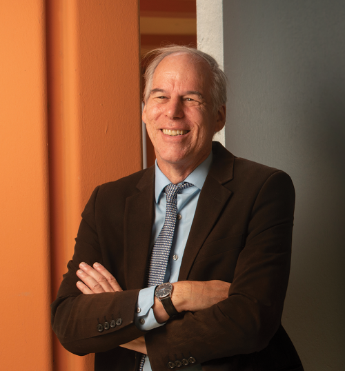 Smiling silver-haired man wearing dark suit jacket, tie and dress shirt, arms crossed, leaning against orange building column