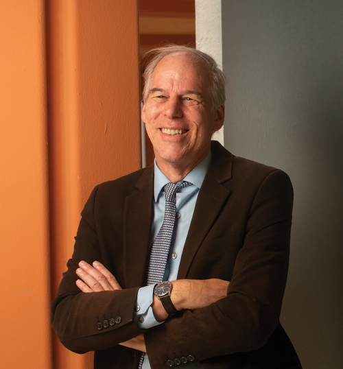 Smiling silver-haired man wearing dark suit jacket, tie and dress shirt, arms crossed, leaning against orange building column 